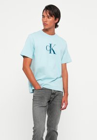 Muškarac nosi svijetloplavu Calvin Klein majicu i sive traperice, stoji s jednom rukom u džepu, gleda sa strane na bijeloj pozadini.