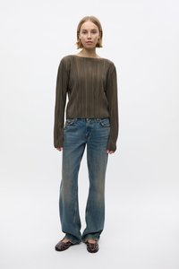Bruine, geribbelde long-sleeve top gecombineerd met vervaagde blauwe jeans. Het model staat met de handen aan de zijkanten op een effen achtergrond.