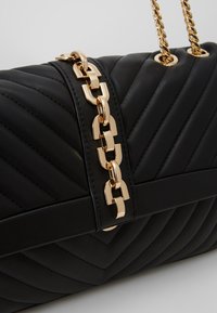 Černá prošívaná kožená taška se zlatým řetězovým popruhem a obdélníkovým zlatým řetězovým akcentem na předním klopě na jednolitém pozadí.