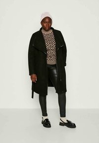 Cappotto lungo nero con un ampio colletto, indossato sopra un maglione beige a stampa e leggings in pelle nera. Il modello lo abbina a scarpe nere chunky.