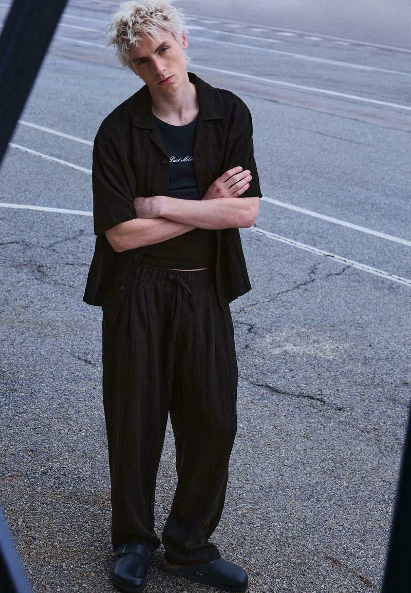 Ung person med kort blekt hår står på asfalt med armarna korsade, klädd i lösa svarta kläder och svarta slip-ons.