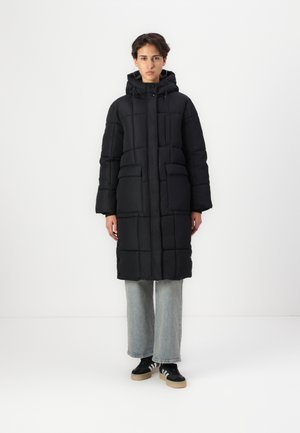 Parka noir rembourrée avec capuche, grandes poches avant et coupe droite, portée sur un jean large bleu clair et des baskets noires.