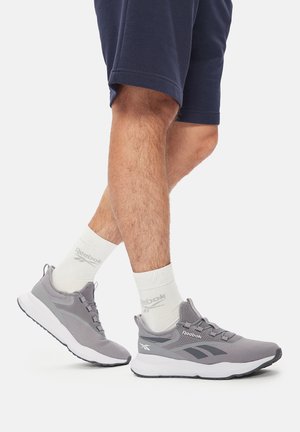 Sivi Reebok tenisice s mrežastim gornjim dijelom, teksturiranim detaljima i bijelim potplatom. Nosene s bijelim čarapama i tamnoplave kratke hlače, otkrivajući atletske listove.