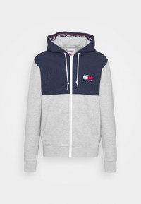 Sudadera con capucha que presenta una sección superior en azul marino y una sección inferior en gris claro, con cremallera frontal y bolsillos laterales. Logo "TOMMY JEANS" en el pecho.