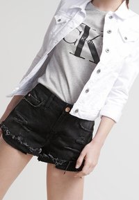 Osoba oblečená v bílé zapínací bundě, šedém tričku s logem Calvin Klein a černých džínových šortkách s obroušenými okraji.