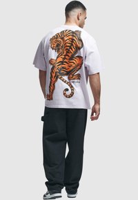 Hvit bomulls t-skjorte med et stort oransje og svart tigermotiv på ryggen. Modellen har på seg svarte, løstsittende bukser og sneakers.