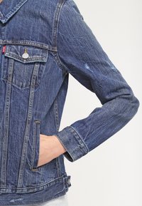 Denim jacket in dark blue, featuring a classic design, two front pockets, and distressed details on the sleeves. Silver button accents.