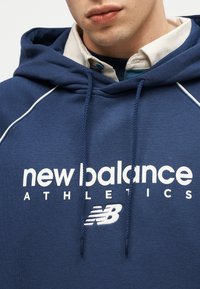 Close-up van een persoon die een blauw New Balance Athletics-hoodie draagt met wit logo en trekkoorden, over een overhemd met kraag.