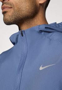 Μπλέ φανέλα με φερμουάρ και ψηλό γιακά, με μικρό ασημένιο λογότυπο Nike στο στήθος και ματ υφή.