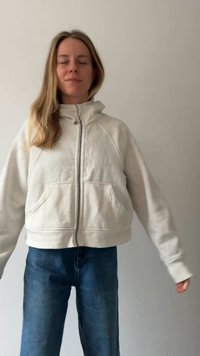 Sudadera corta beige claro con cremallera, cuello alto, bolsillos frontales y mangas largas. Combinada con vaqueros azul oscuro. Textura suave.