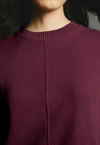 Burgundowa dziergana sweter z prążkowanym okrągłym dekoltem i detalem szwu z przodu, charakteryzująca się miękką teksturą i średniej wagi.