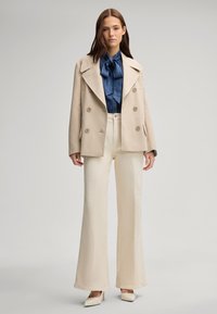 Beige double-breasted jas over een blauwe blouse met een strikje, gecombineerd met crèmekleurige wijde broek en puntige beige hakken.