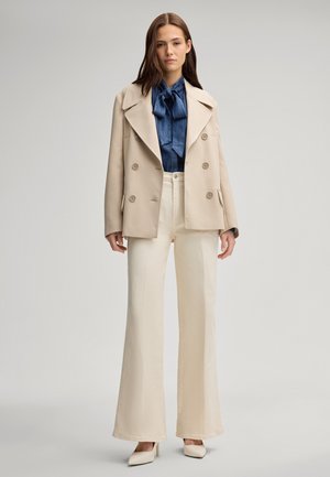Cappotto beige doppiopetto sopra una blusa blu con fiocco, abbinato a pantaloni crema a gamba larga e décolleté beige a punta.