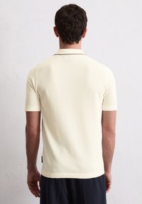 Koszulka polo z krótkim rękawem w jasnej kremowej barwie, o prążkowanej fakturze, z klasycznym kołnierzykiem i kontrastowymi wykończeniami. Widok z tyłu ukazuje jej dopasowany krój.