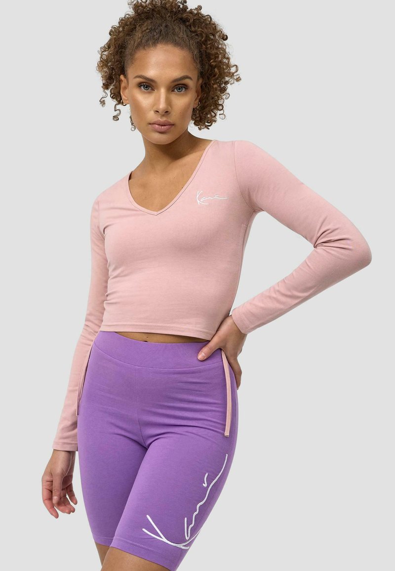 Rosa langermet crop topp med V-hals og hvit logo. Kombinert med lilla høyt liv sykkelshorts med hvit linjedetalj.