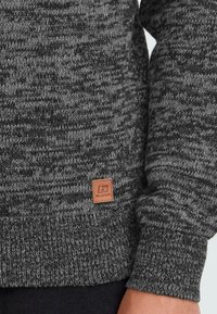 Primo piano del torso e del braccio di una persona che indossa un maglione lavorato a maglia grigio scuro con una piccola etichetta in pelle marrone con il logo Blend.