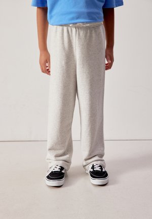 Vaikas dėvintis šviesiai pilkus sportinius kelnes ir juodai baltus sportinius batelius, stovintis priešais paprastą šviesų foną.