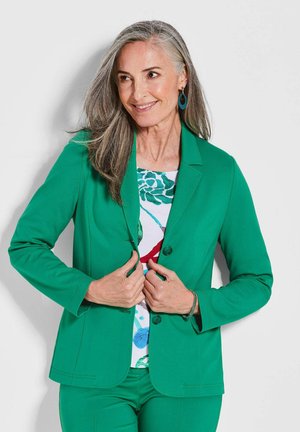 Grüner maßgeschneiderter Blazer mit glatter Textur, Ein-Knopf-Verschluss und eingezogenen Revers; getragen über einem blumengemusterten Oberteil.