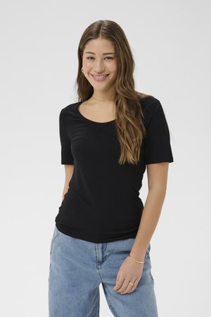 Mlada žena s dugom smeđom kosom, odjevena u crnu kratku majicu i svijetloplave traperice, smiješi se i stoji ispred jednobojne pozadine.