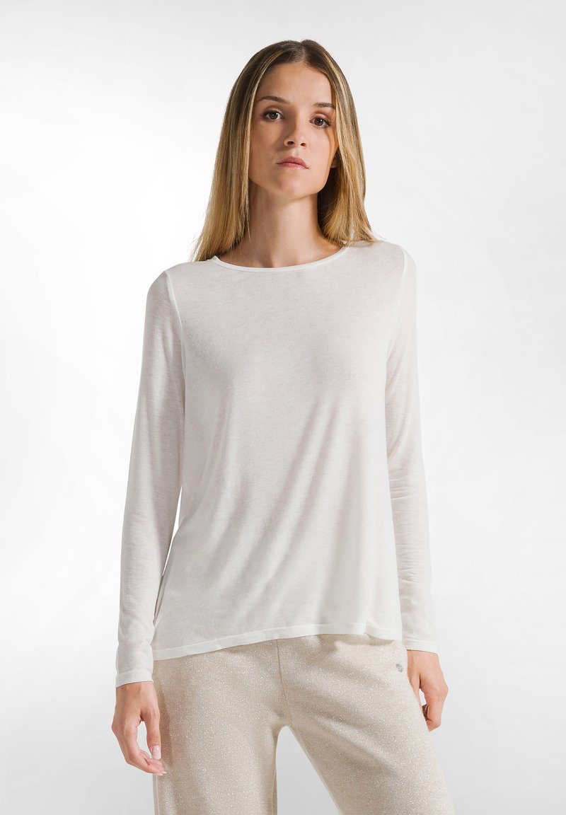 Maglietta bianca a maniche lunghe con scollo rotondo, realizzata in un tessuto sottile e testurizzato, dal taglio morbido, indossata con pantaloni chiari e brillanti.