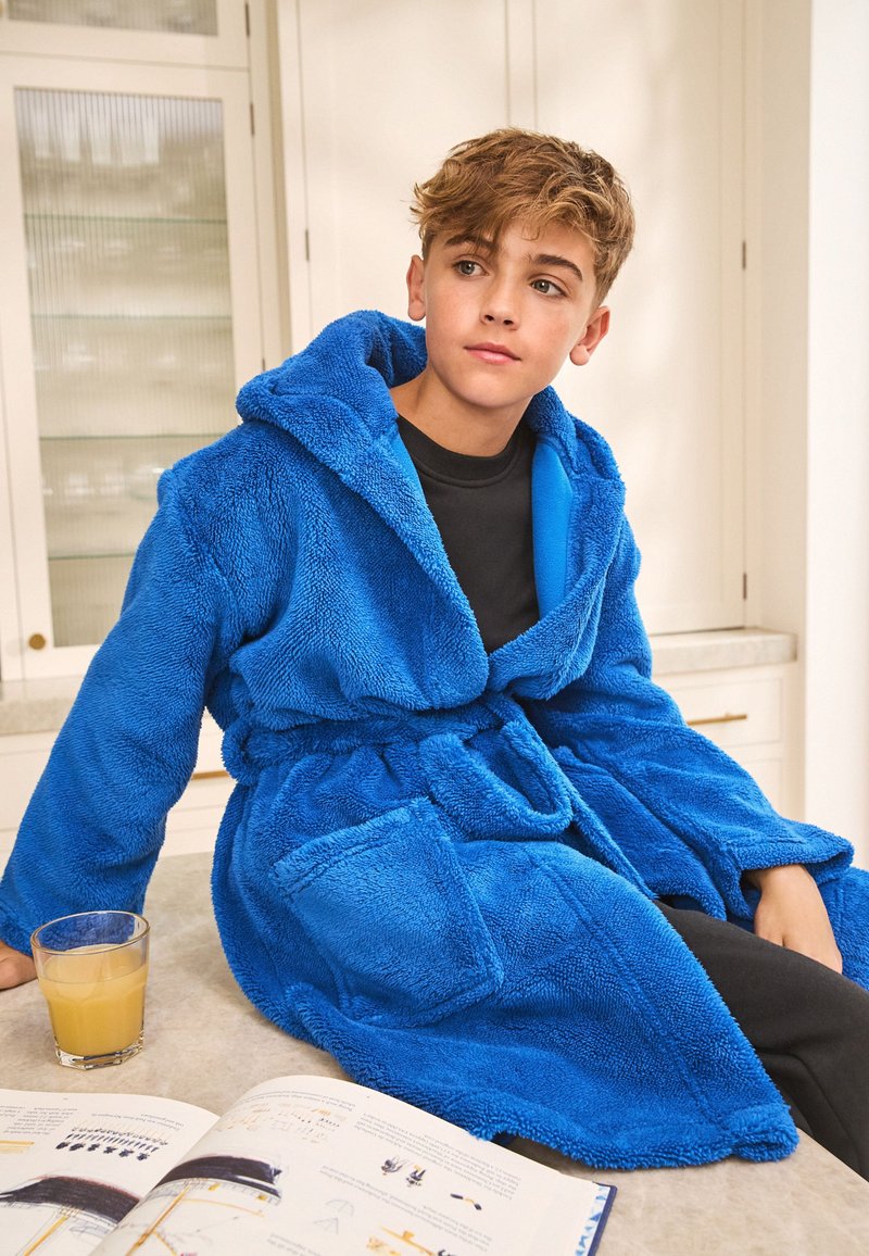 Peignoir à capuche bleu en matière pelucheuse, avec ceinture à nouer et poches latérales. Jeune personne assise, avec une boisson et un livre ouvert à proximité.