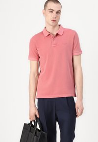 Roze katoenen polo met kraag en korte mouwen, gecombineerd met marineblauwe broek, vastgehouden met een zwart leren tote bag.