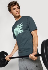 Herre charcoal grå bomulds T-shirt med mintgrøn "Nike" logo og penselstræksdesign, parret med grå sweatpants, mens der holdes en sort vægtstang.