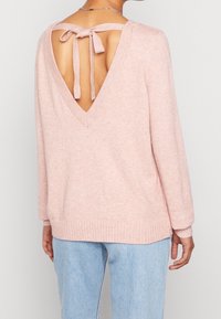 Rosa tröja med djup V-öppning bak, knuten med matchande tyg, ribbade muddar och nederkant, mjuk textur, bärs över ljusblå jeans.