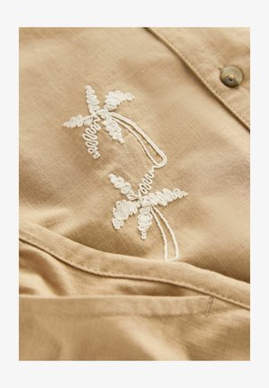Beige linen shirt featuring white embroidered floral patterns, smooth texture, and a button closure. Detailed stitching accentuates design.