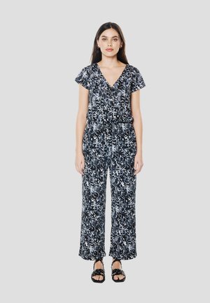 Sort og hvid mønstret jumpsuit med V-udskæring, korte ærmer og vide ben; kombineret med sorte flade sandaler.