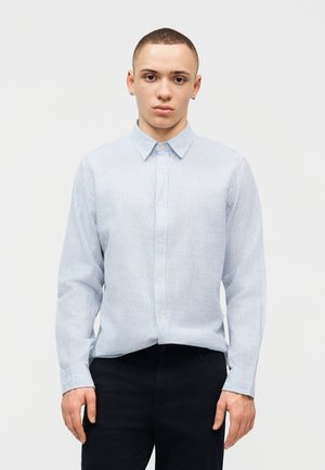 Νεαρός με κοντά μαλλιά που φοράει ένα γαλάζιο πουκάμισο με ρίγες και μαύρο παντελόνι, στέκεται μπροστά σε έναν απλό λευκό φόντο.