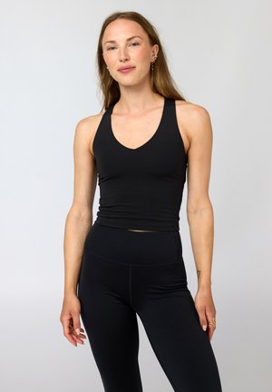 Žena s dlhými vlasmi, oblečená v čiernom tielku bez rukávov a čiernych legínach, stojaca pred jednoduchým svetlým pozadím.