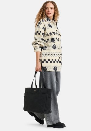 Zwarte totebag met dubbele handvatten, een gladde textuur, een voorzak en een gouden accent. Draagt met een gestreept crèmekleurig shirt en grijze wijde broek.