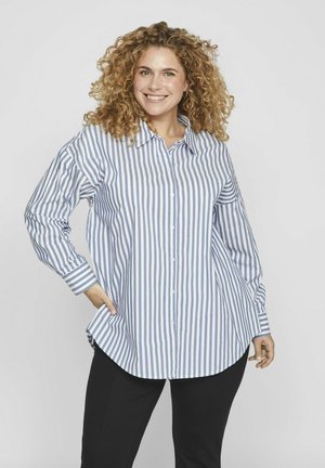 Camisa abotonada azul y blanca a rayas con cuello, manga larga y dobladillo redondeado. Lleva puesta sobre pantalones negros, mostrando un ajuste relajado.
