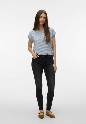 Młoda kobieta stojąca z ręką w kieszeni, ubrana w jasno niebieską koszulkę w paski, czarne obcisłe dżinsy oraz brązowe płaskie buty na białym tle.