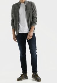 Harmaa neulottu vetoketjullinen neule takki valkoisen t-paidan päällä, yhdistetty tummansinisiin farkkuihin ja harmaa/vihreisiin lenkkareihin. Minimalistinen, istuva malli.