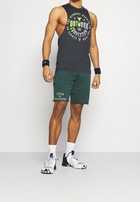 Camiseta sin mangas oscura con texto brillante, pantalones cortos verdes con la impresión "IRON PARADISE" y zapatillas de deporte blancas con acentos negros.