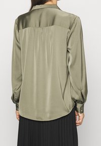 Chemise en satin vert olive avec un pli au dos, manches longues et poignets boutonnés, dotée d'un ourlet arrondi et d'une texture lisse.