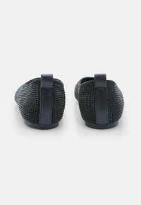 Pár čiernych ponožkových plochých topánok s textúrovaným zvrškom a vertikálnym ťahacím poutkom, pohľad zozadu na bielom pozadí.