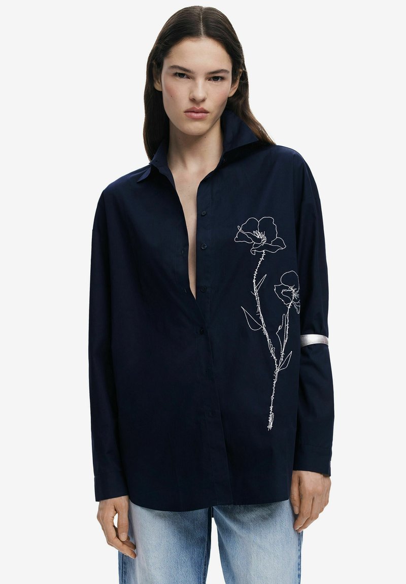 Marineblaue Hemdbluse mit hohem Kragen und langen Ärmeln. Mit weißen floralem Stickereien auf der Vorderseite und einem silbernen Akzent am Ärmel.