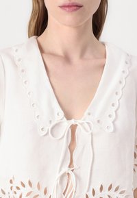 Hvit bluse med en scalloped krage med hullbroderi, knyting i halsen og broderte utskjæringer i kanten.