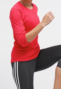 Sportowa bluzka z długim rękawem w jaskrawoczerwonym kolorze, wykonana z teksturowanej tkaniny, zestawiona z czarnymi legginsami ozdobionymi białymi paskami po bokach, w aktywnej pozie.