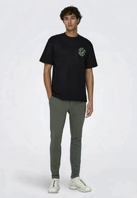 T-shirt de algodão preto com gráfico circular verde no peito, combinado com calças justas em verde-oliva e sapatilhas brancas.