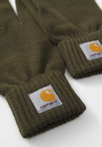 Grønne strikkede handsker med ribbede manchetter, med et kvadratisk mærke med et gult logo og "Carhartt" tekst. Blød tekstur og afslappet design.