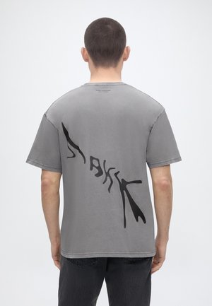 Άνδρας με κοντά μαλλιά φοράει γκρι μπλουζάκι με μαύρο αφηρημένο διαγώνιο κείμενο και μαύρο παντελόνι, στέκεται μπροστά σε απλό φωτεινό φόντο.