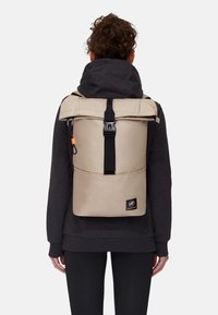 Beige rulltop ryggsäck med svart spänne. Har en framficka och logotypapplikation. Texturerad tyg och justerbara remmar.