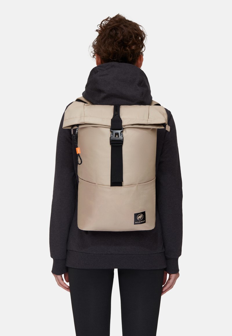 Beige rulltop ryggsäck med svart spänne. Har en framficka och logotypapplikation. Texturerad tyg och justerbara remmar.