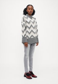 Maglione a maglia grigio con motivo a chevron e colletto alto, abbinato a jeans skinny grigio chiaro e scarpe con tacco rosse lucide.