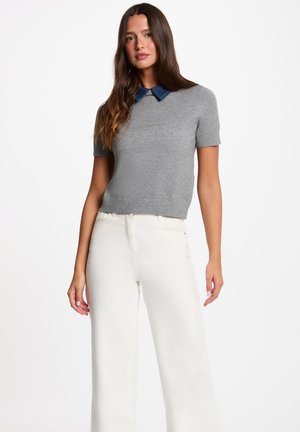 Mujer con un suéter gris de manga corta con cuello azul oscuro y pantalones anchos blancos de talle alto, de pie frente a un fondo liso.