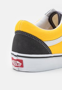 Gul canvas og svart semsket skinn sneakers med hvit søm og detaljer, som har en rød Vans-logo på hælen og en teksturert gummisåle.
